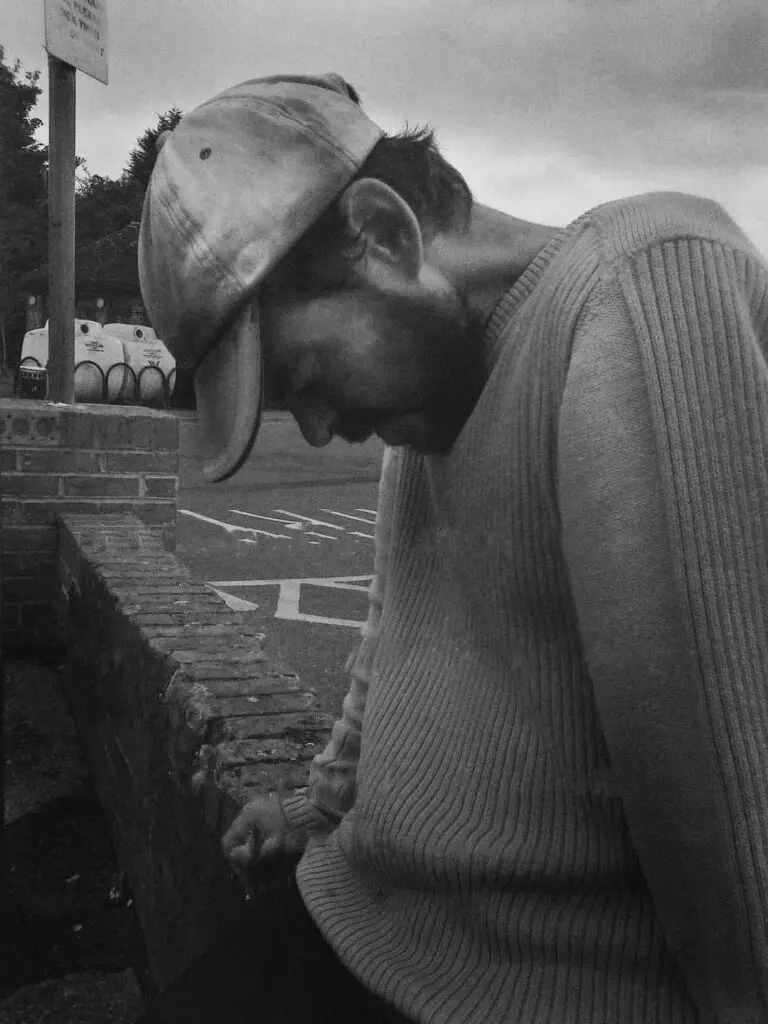 A man in a worn jumper and cap sitting against a low brick wall, head bowed, in a quiet outdoor setting