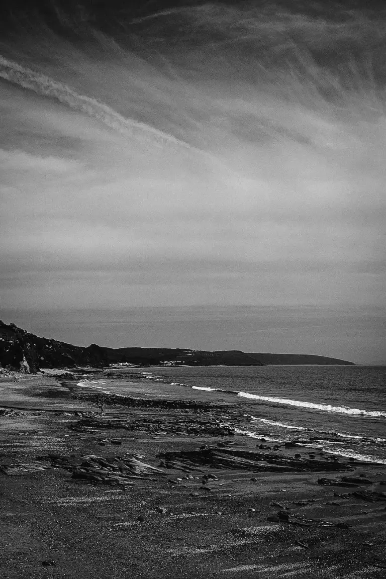 Black and white photograph of a rocky beach shoreline with gentle waves and a dramatic, cloud-streaked sky