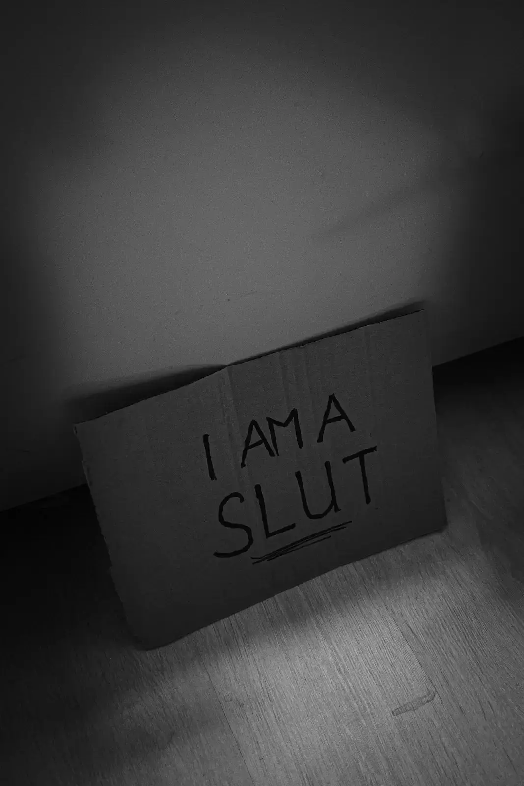 A cardboard sign on the floor with the handwritten words “I AM A SLUT,” lit by a single spotlight in an otherwise shadowed room