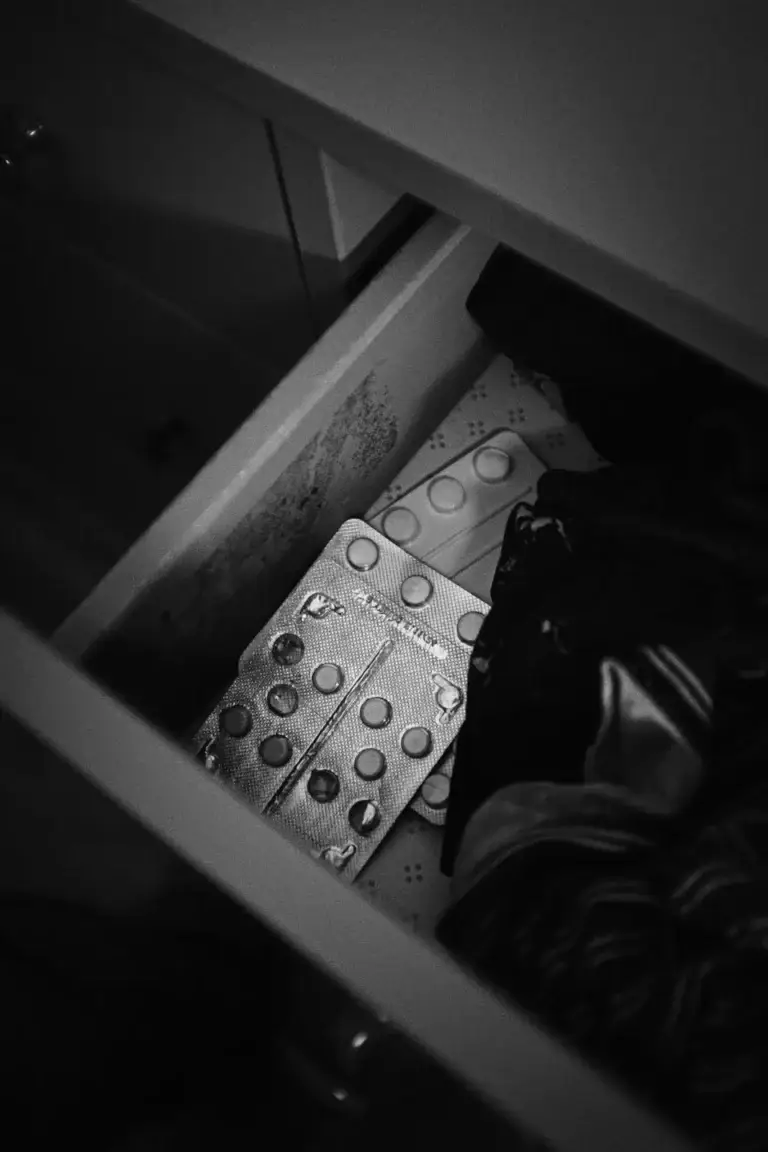 A black and white photo of a half-open drawer containing blister packs of medication and clothes