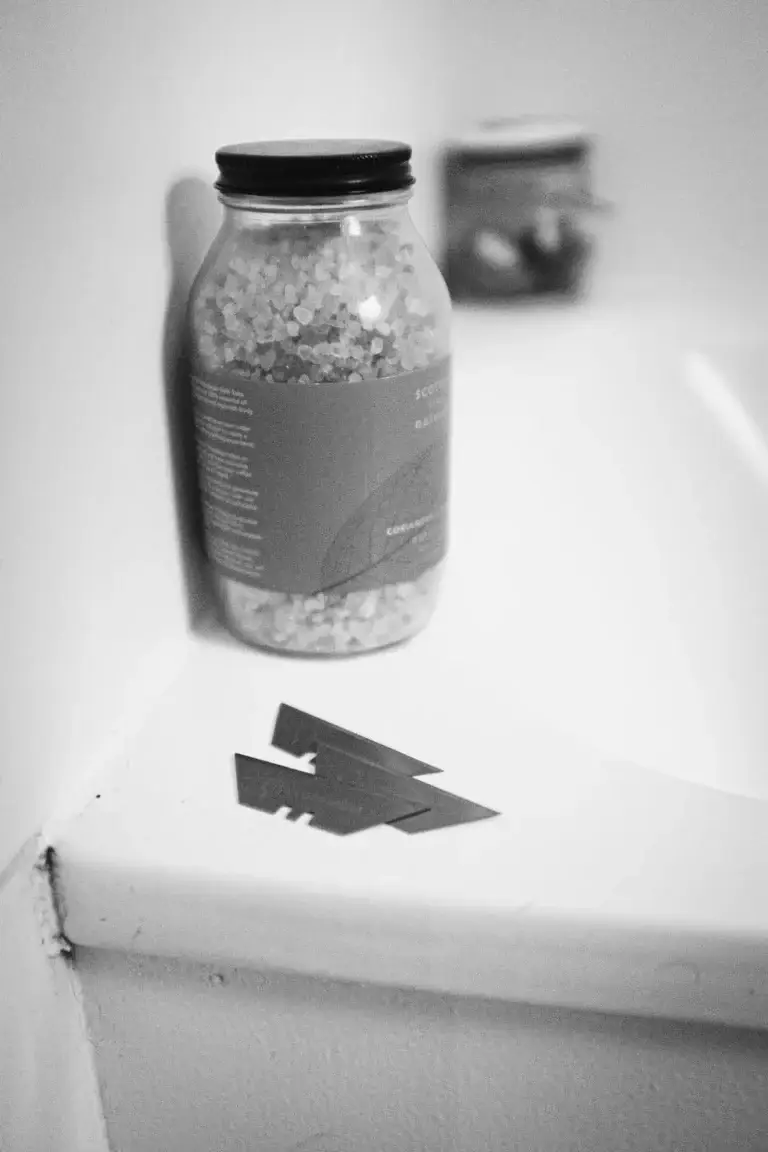 A black and white image of a jar of bath salts placed beside two razor blades on the edge of a bath