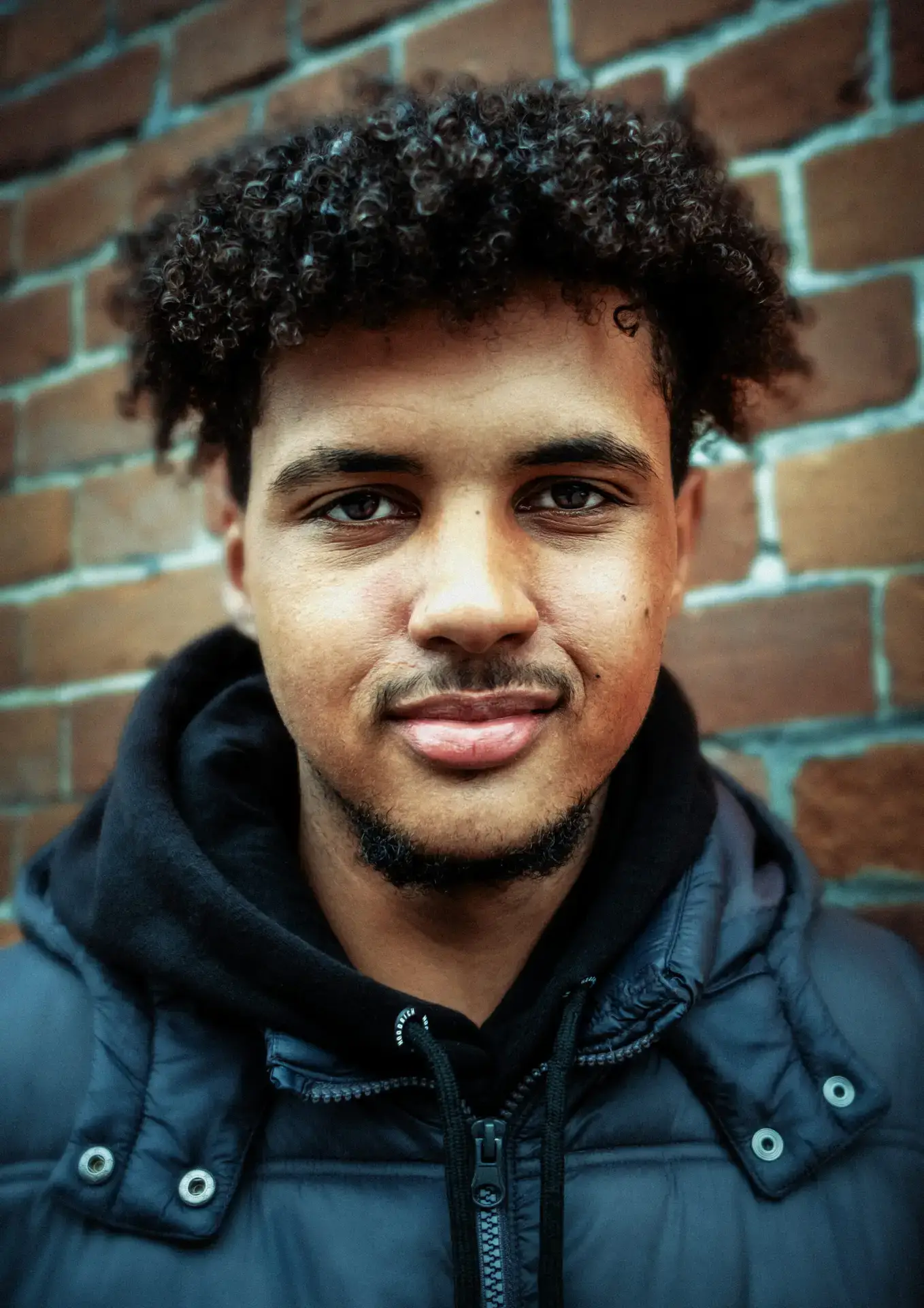 Portrait of a young man with curly hair and a dark padded jacket, standing confidently in front of a brick wall