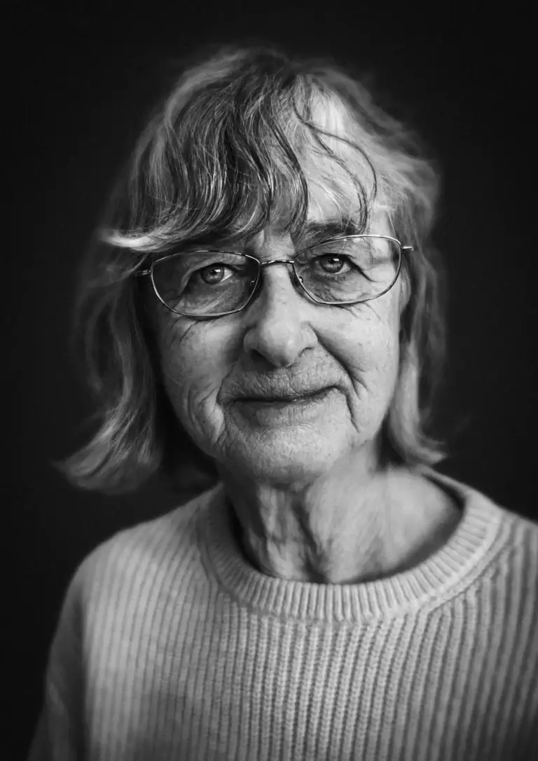 Black and white portrait of an older woman with shoulder-length hair and glasses, wearing a ribbed jumper and a calm expression