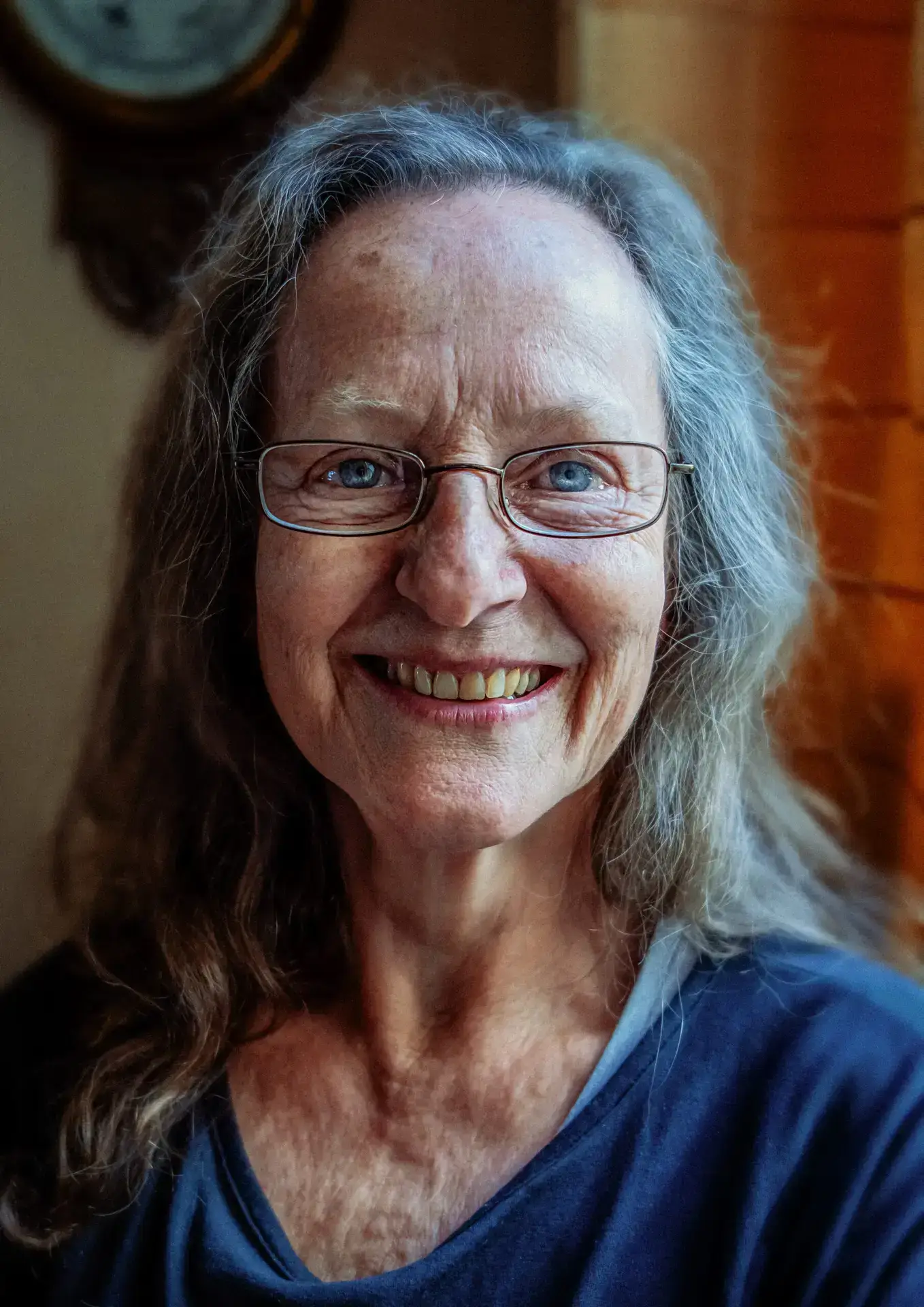 Portrait of an older woman with long grey hair and glasses, smiling warmly with light reflecting in her blue eyes