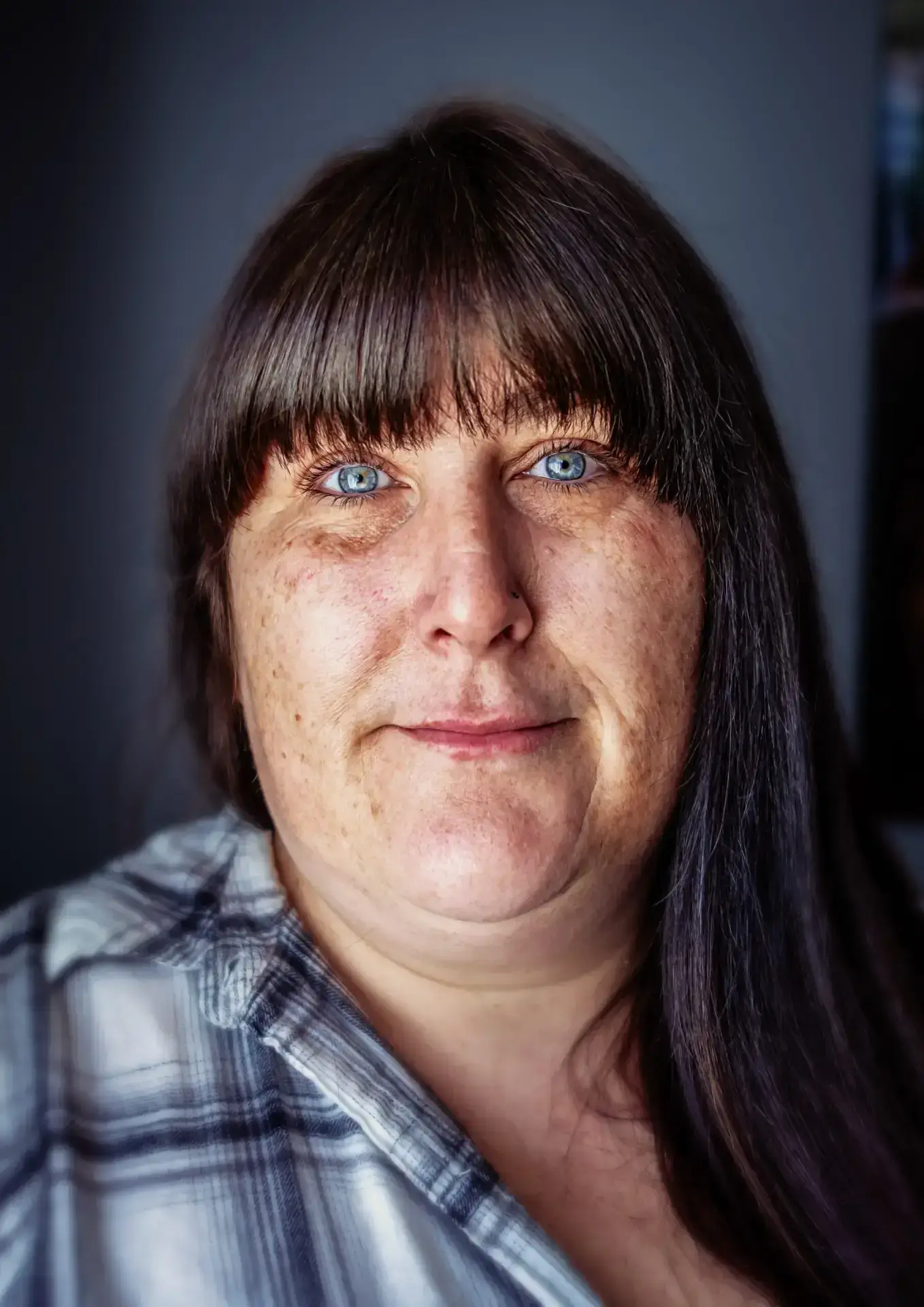 Close-up portrait of a woman with long dark hair and striking blue eyes, wearing a plaid shirt and looking directly at the camera