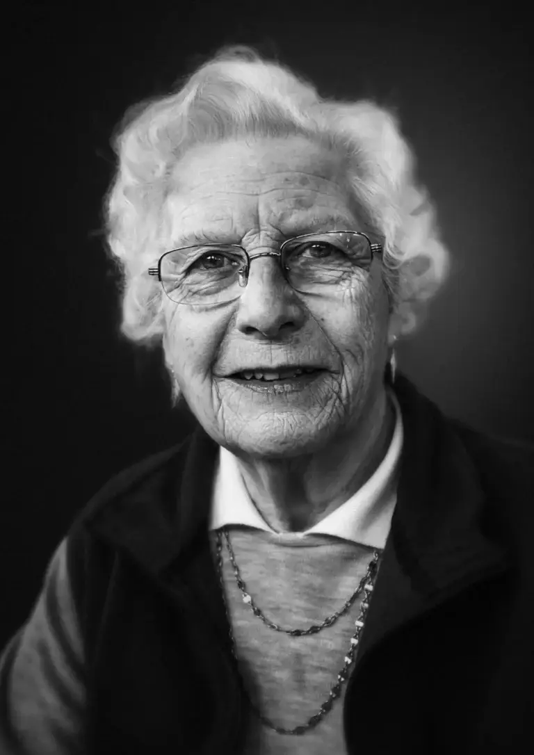Black and white portrait of an elderly woman with white hair, glasses, and a soft smile, wearing layered clothing and jewellery