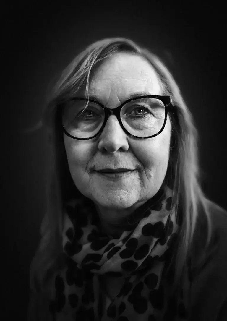 Black and white portrait of an older woman with glasses and a patterned scarf, softly lit against a dark background
