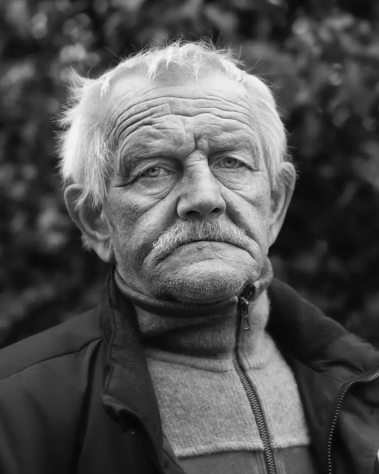 Black-and-white close-up portrait of an elderly man with white hair and a moustache, wearing a fleece and jacket, looking directly at the camera.