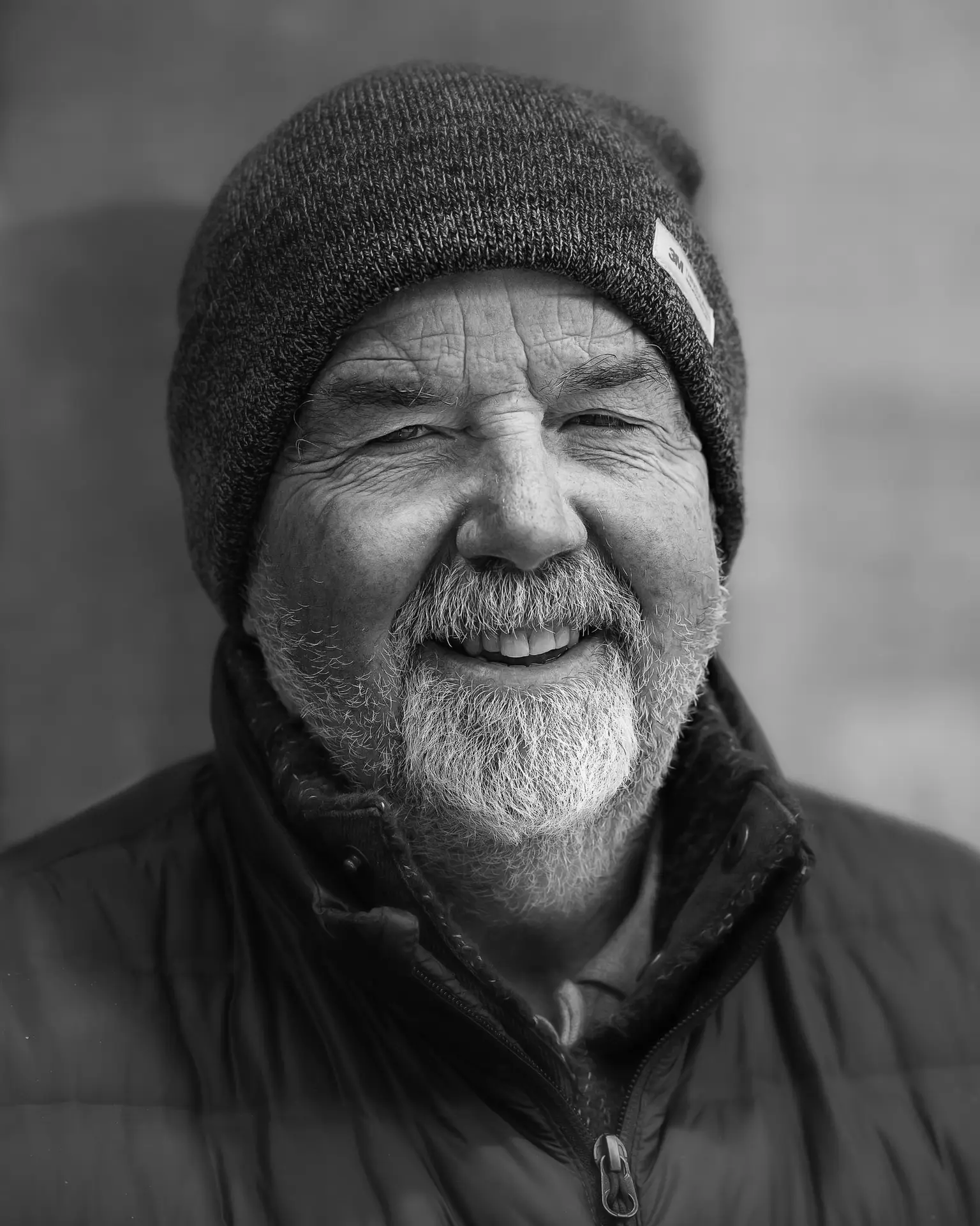 Black and white portrait of an older man with a beanie hat and a warm padded jacket, smiling warmly at the camera