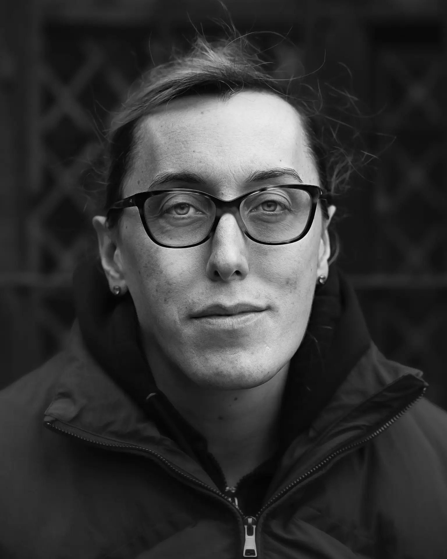 Black and white portrait of a person wearing glasses and a zipped jacket, looking straight at the camera with a calm and steady expression