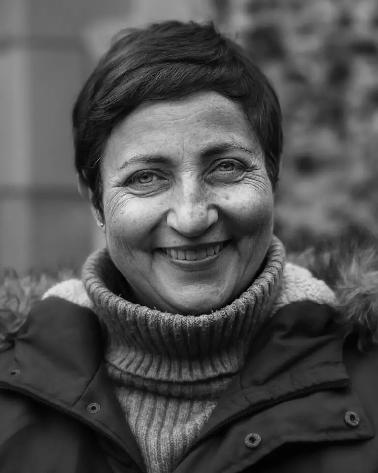 Black-and-white close-up portrait of a smiling woman with short dark hair, wearing a turtleneck jumper and winter coat with a fur-trimmed hood.