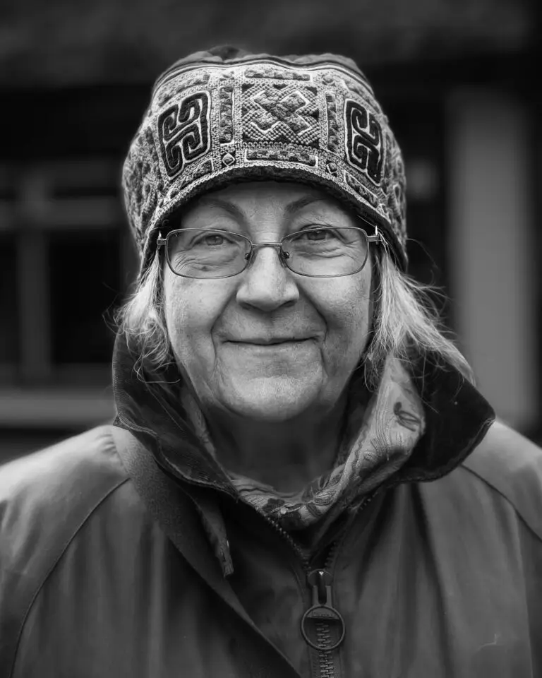 Black-and-white close-up portrait of an older woman wearing glasses, a patterned winter hat, and a zipped coat, smiling softly at the camera.