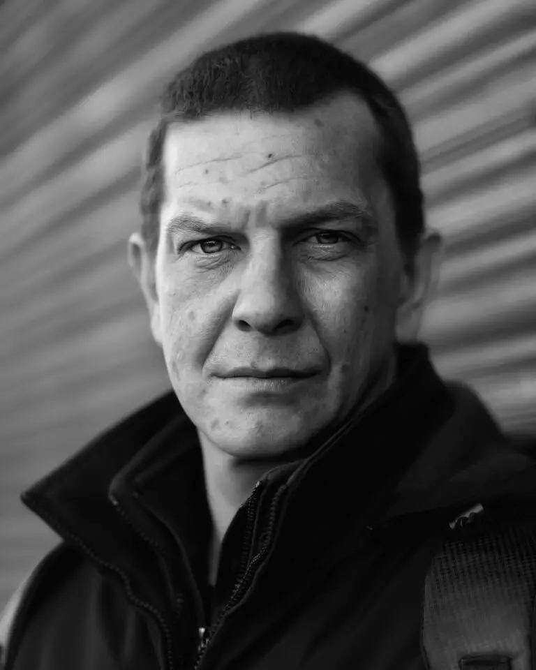 Black-and-white close-up portrait of a man with short hair, wearing a zipped jacket, standing in front of a blurred metal shutter and looking directly at the camera.