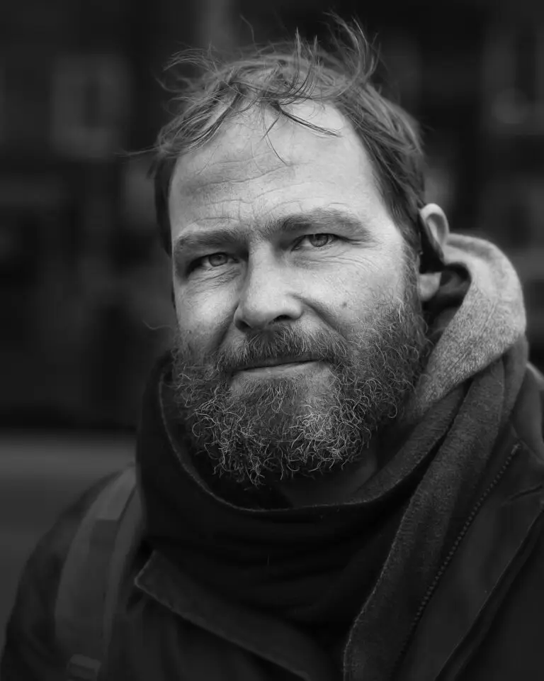Black-and-white close-up portrait of a bearded man with tousled hair, wearing layered winter clothing and looking directly at the camera.