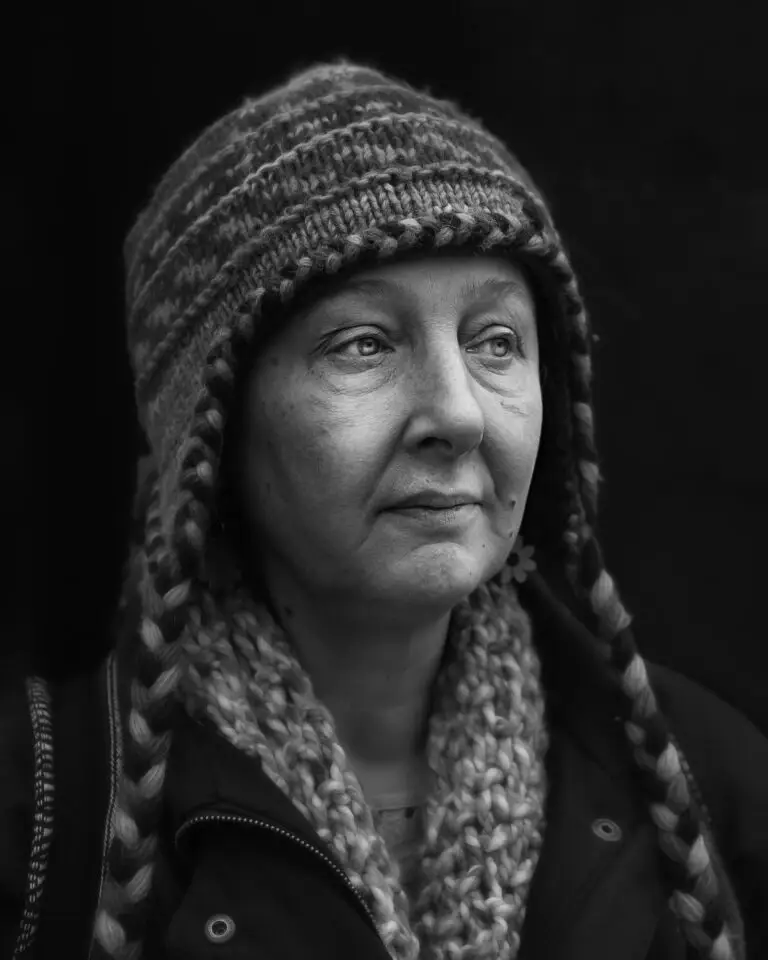 Black-and-white close-up portrait of a woman wearing a knitted hat with ear flaps and a chunky scarf, looking thoughtfully into the distance.