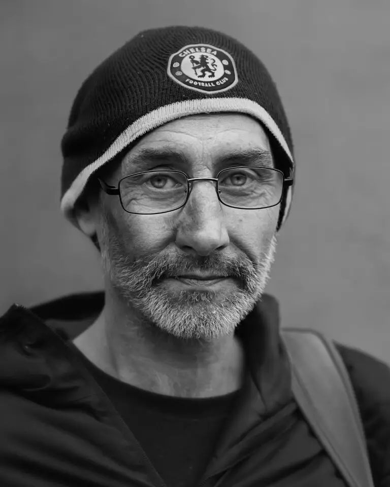 Black-and-white close-up portrait of a man with glasses and a beard, wearing a Chelsea Football Club beanie and casual jacket, looking at the camera.