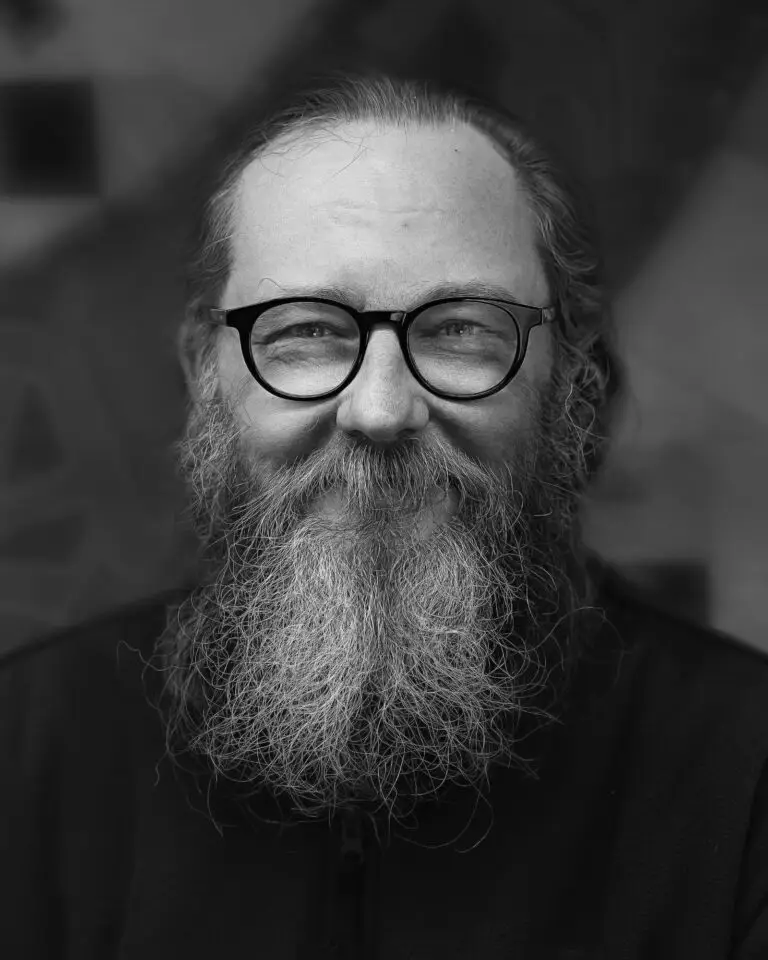 Black-and-white close-up portrait of a bearded man with glasses, smiling warmly at the camera against a blurred background.