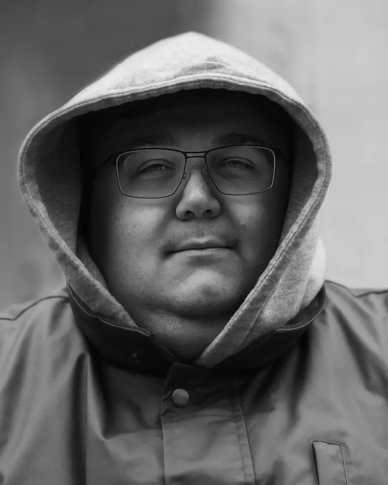 Black-and-white close-up portrait of a man wearing glasses, a hooded sweatshirt, and a buttoned jacket, looking calmly at the camera.