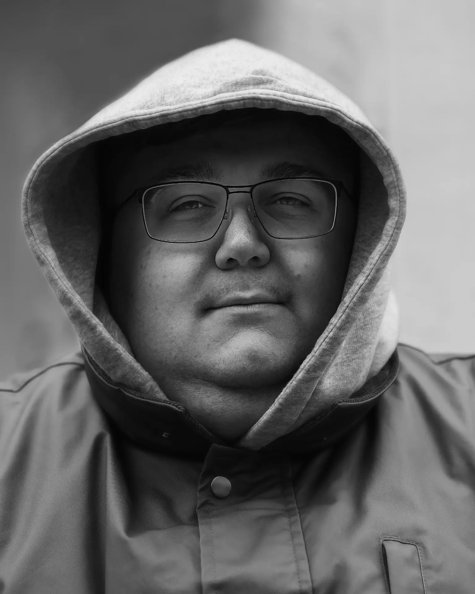 Black-and-white close-up portrait of a man wearing glasses, a hooded sweatshirt, and a buttoned jacket, looking calmly at the camera.