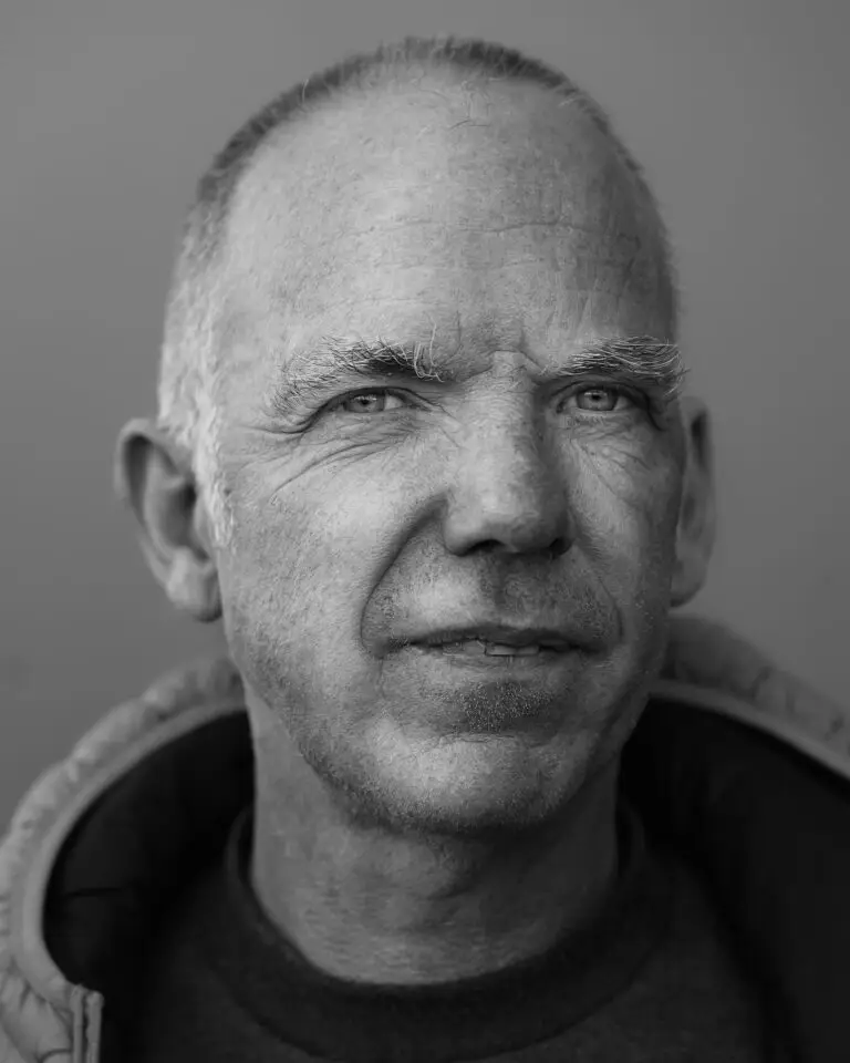 Black-and-white close-up portrait of a man with short-cropped hair, light stubble, and a calm expression, looking slightly past the camera.