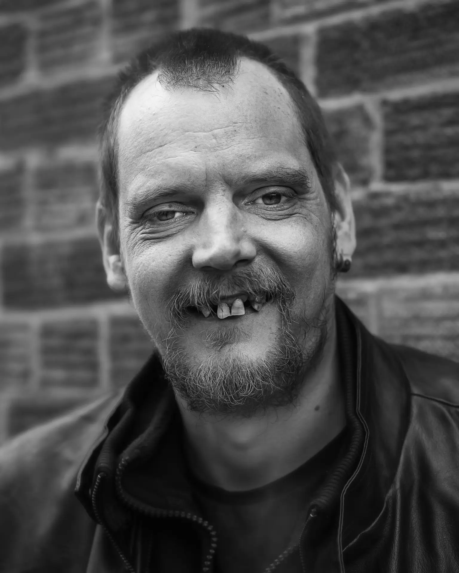 Black and white portrait of a middle-aged man with missing teeth, wearing a leather jacket and smiling warmly