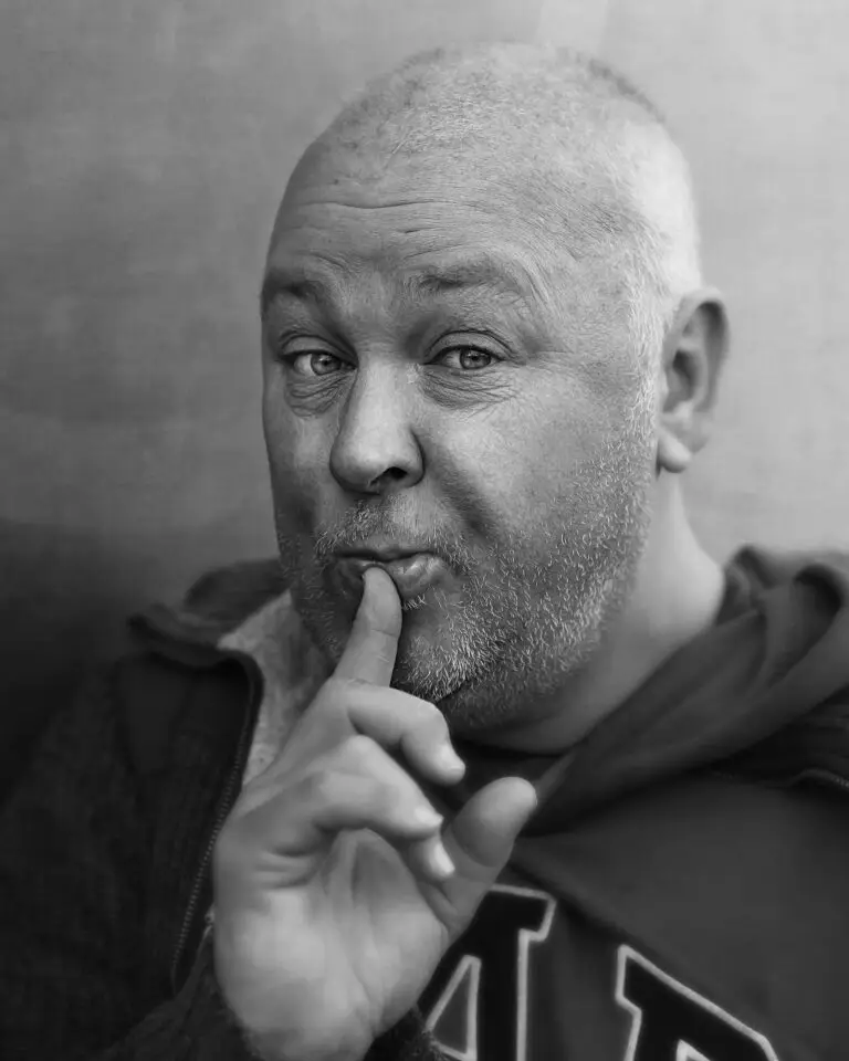 Black and white portrait of a middle-aged man with cropped hair and stubble, holding one finger to his lips with a playful, knowing expression.