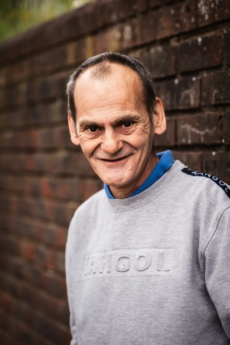 Portrait of a smiling man in a grey Kangol sweatshirt, standing in front of a brick wall