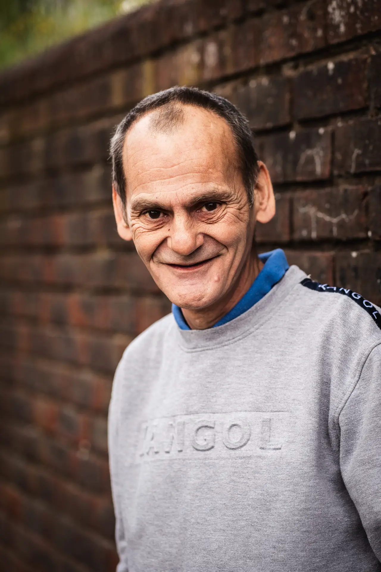 Portrait of a smiling man in a grey Kangol sweatshirt, standing in front of a brick wall