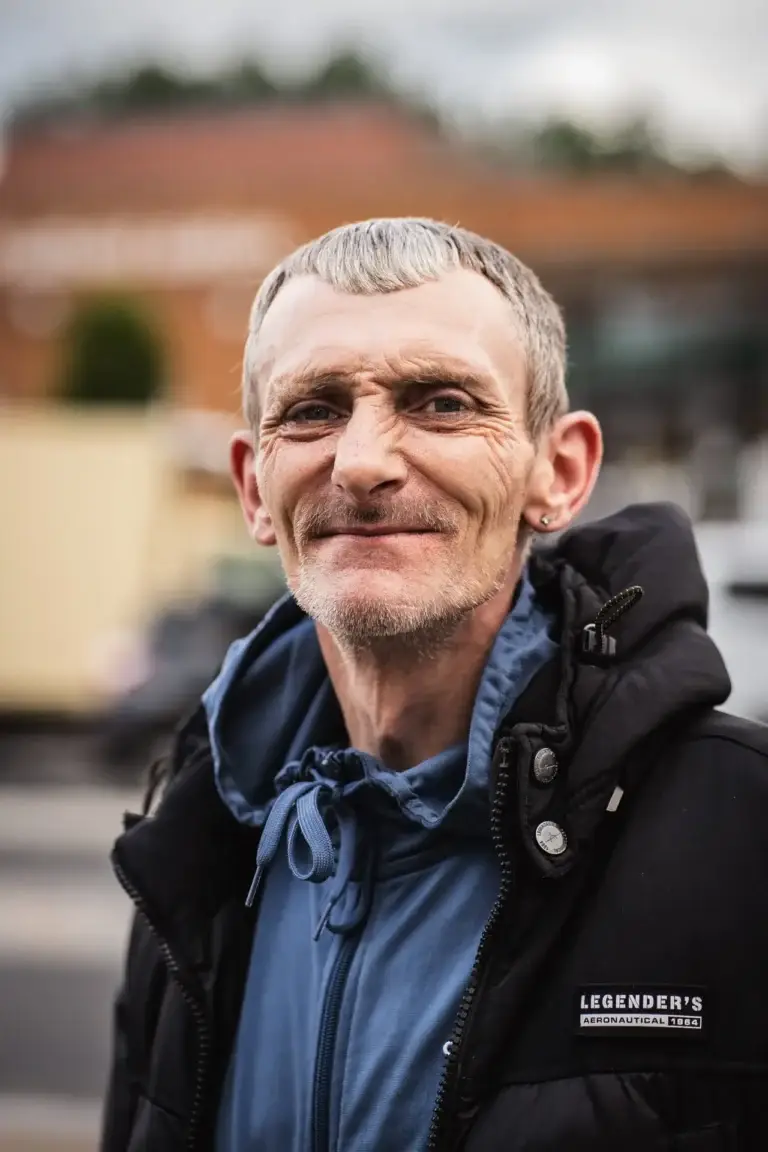 homeless 6 Portrait of a man in a blue hoodie and black coat, smiling slightly while standing outdoors