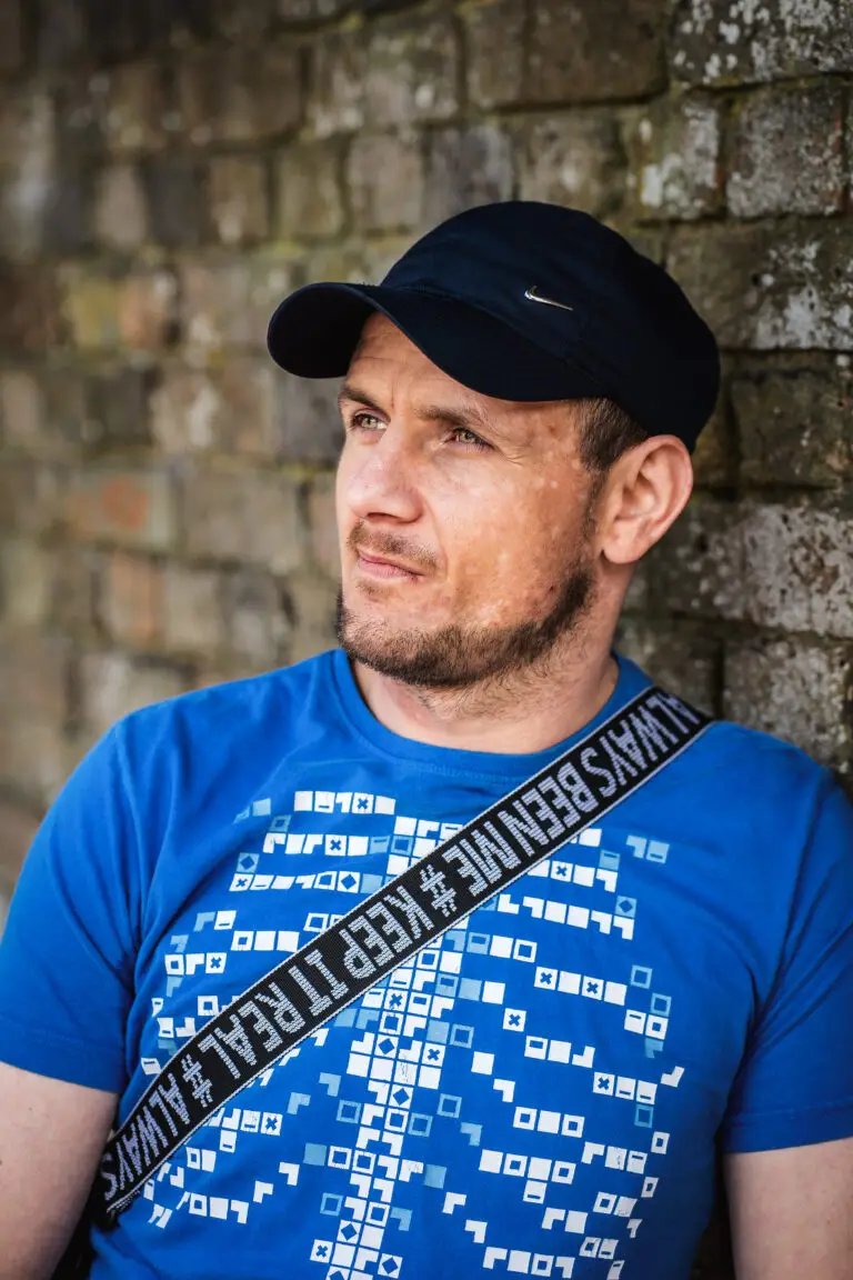 Man in a blue graphic t-shirt and black cap looking thoughtfully to the side while leaning against a brick wall