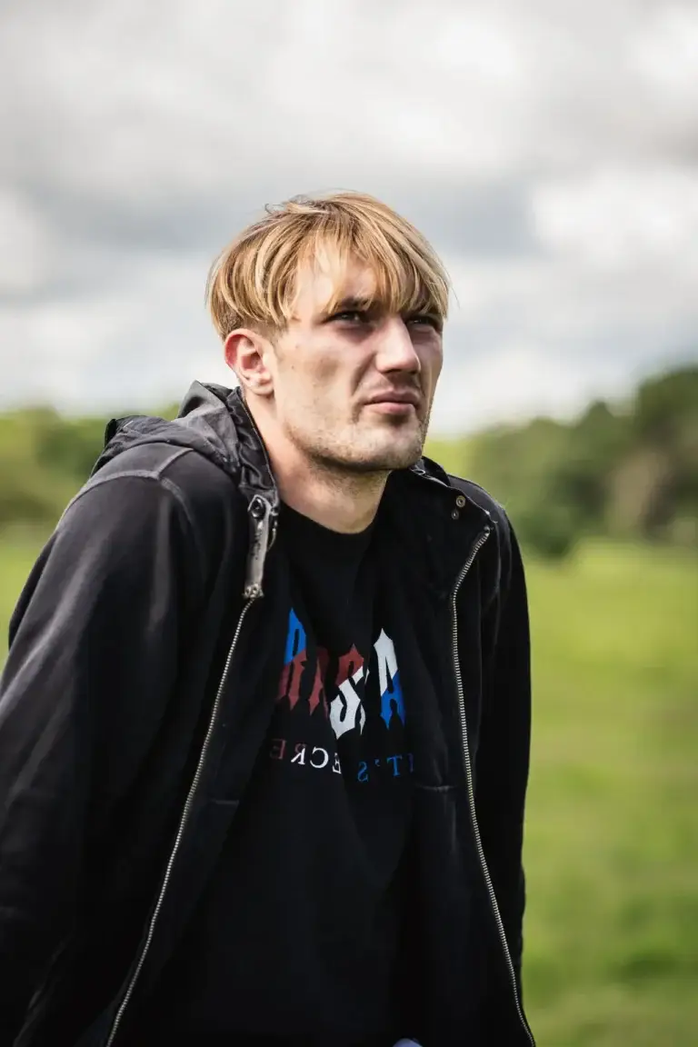 Young man with blonde hair in a black hoodie standing in a grassy field, looking into the distance with a tense expression