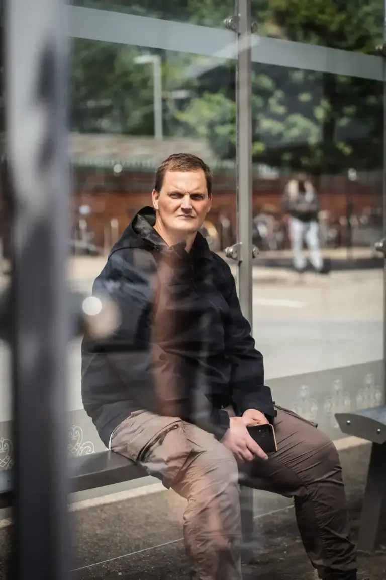 Man sitting in a glass bus shelter holding a phone, wearing a black jacket and beige trousers, looking out with a thoughtful expression