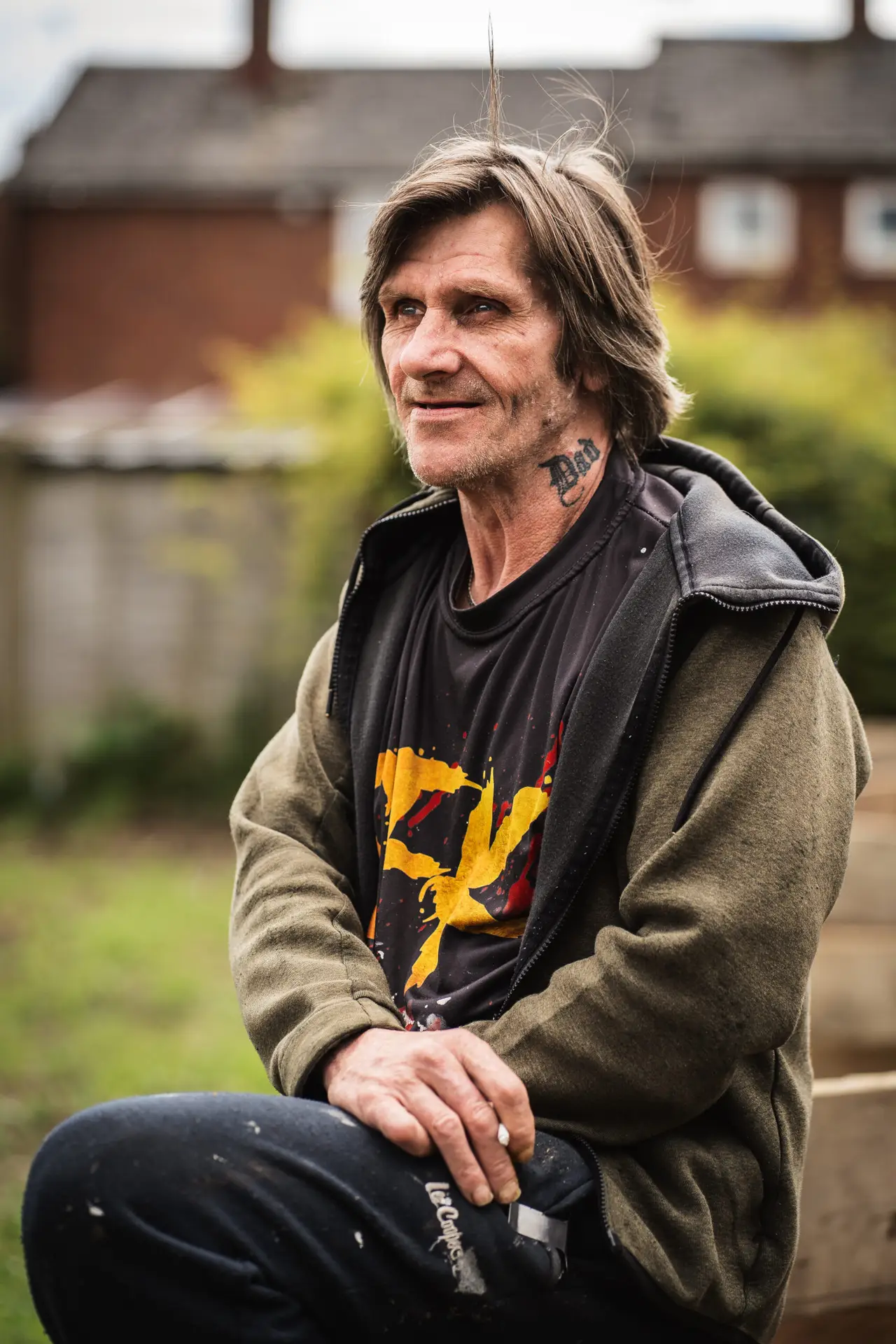 Colour portrait of a man with a “Dad” tattoo on his neck, wearing a hoodie and graphic T-shirt, sitting outdoors and looking thoughtfully into the distance.