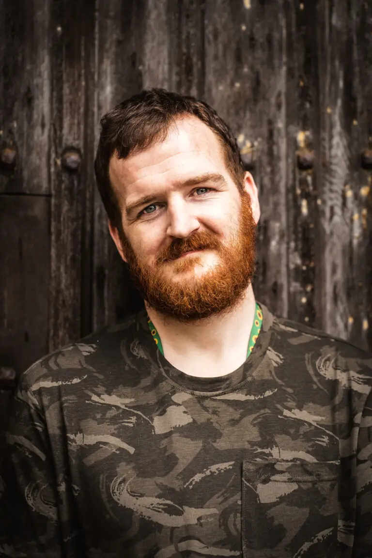 Colour portrait of a man with a red beard and short hair, wearing a camouflage-patterned shirt, smiling gently against a weathered wooden background.