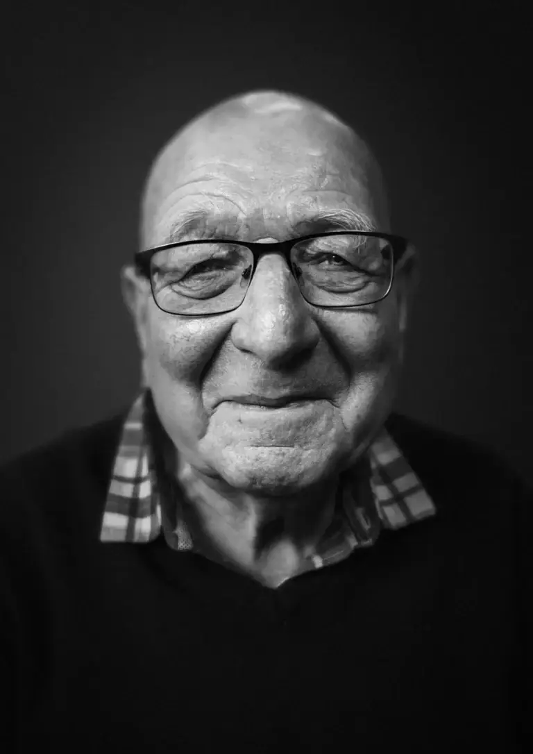 black and white portrait of an elderly man with glasses, a checked shirt under a jumper, and a gentle, closed-lip smile. His expression is warm and content, his eyes kind and full of life.