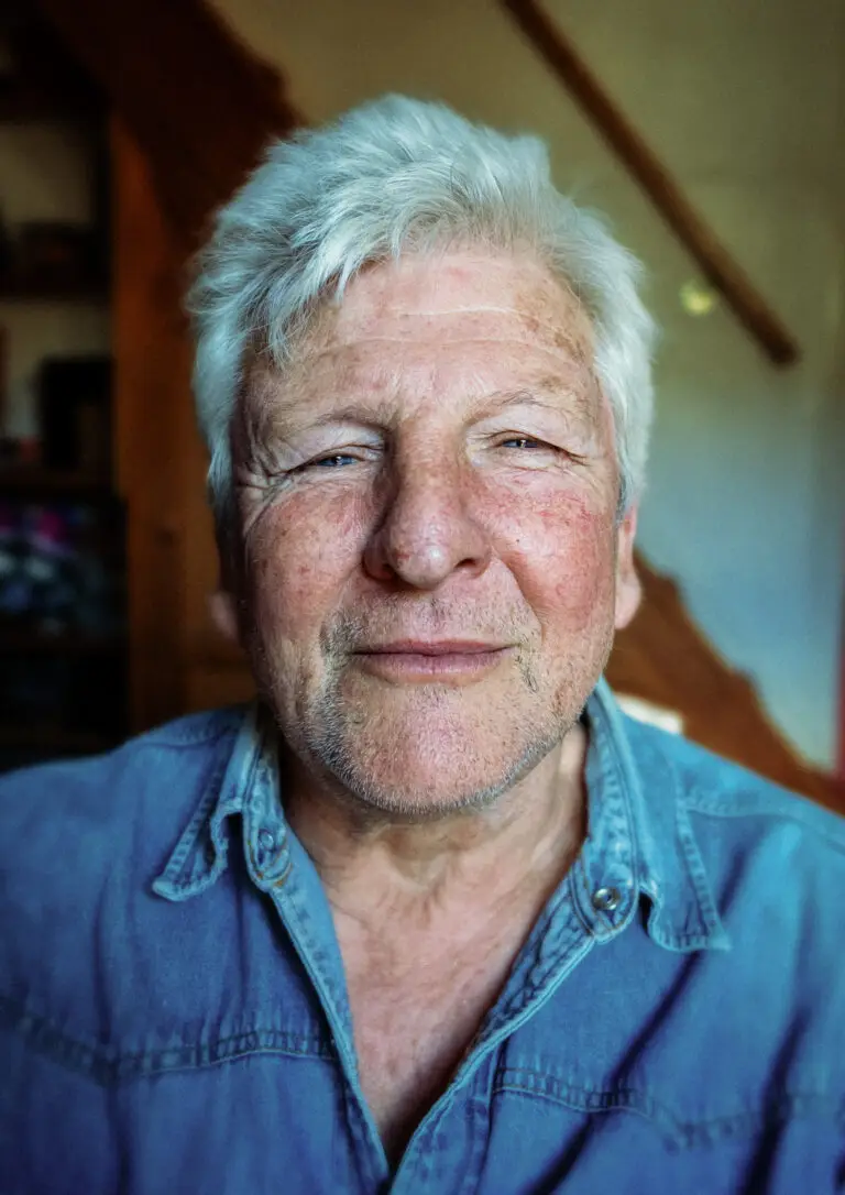Portrait of an older man with white hair and a weathered face, wearing a denim shirt, looking calmly into the camera with a faint smile.