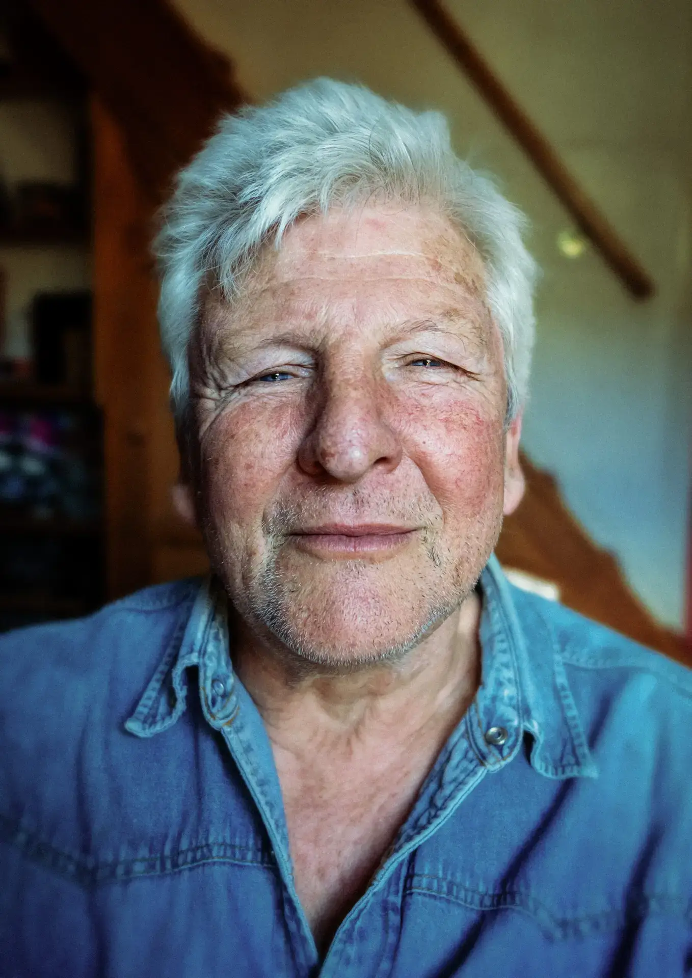Portrait of an older man with white hair and a weathered face, wearing a denim shirt, looking calmly into the camera with a faint smile.