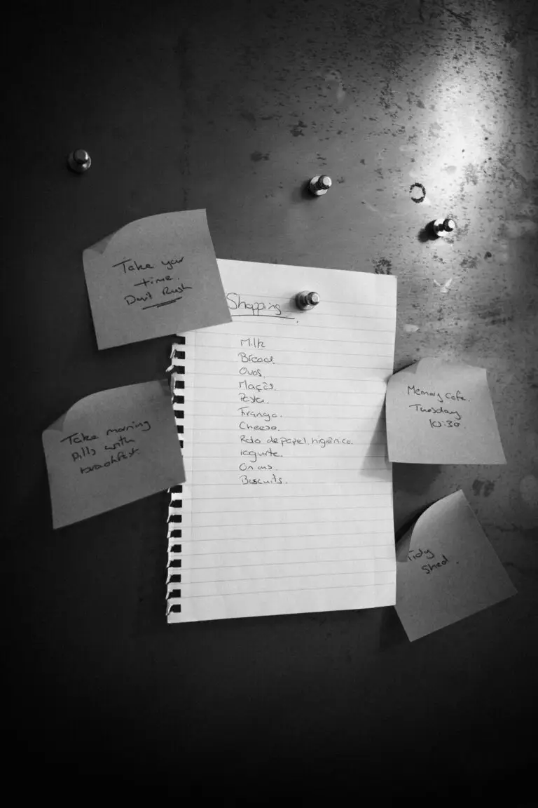 A fridge or noticeboard covered with handwritten reminders: a shopping list on lined paper and Post-it notes saying “Take your time, don’t rush,” “Take morning pills with breakfast,” “Memory café Tuesday 10:30,” and “Tidy shed.”