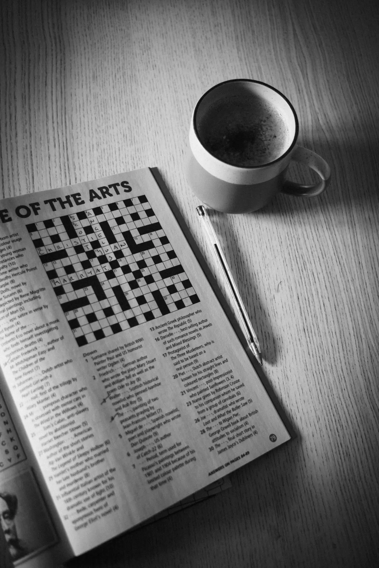 A close-up black and white image of a completed crossword puzzle, a ballpoint pen, and a mug of coffee resting on a wooden table — evoking calm, routine, and quiet concentration.