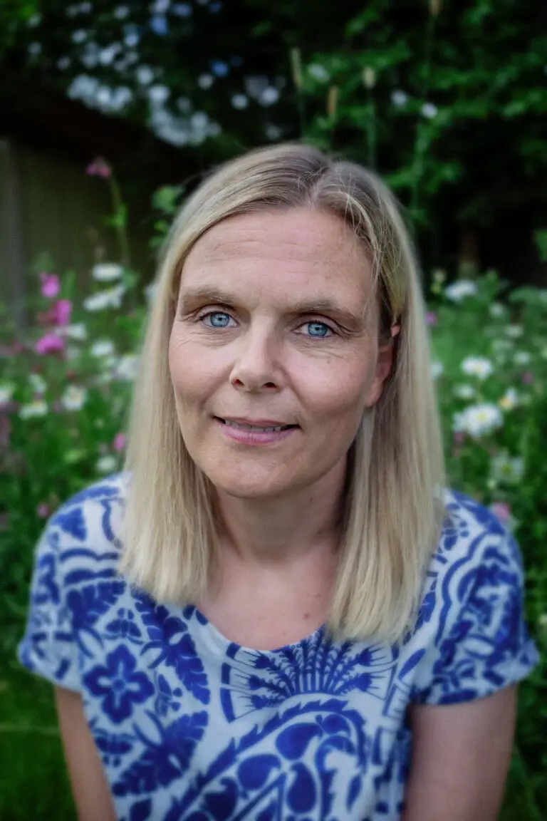 A woman with straight blonde hair and clear blue eyes, wearing a patterned blue and white top, seated outdoors in front of a background of wildflowers and greenery.