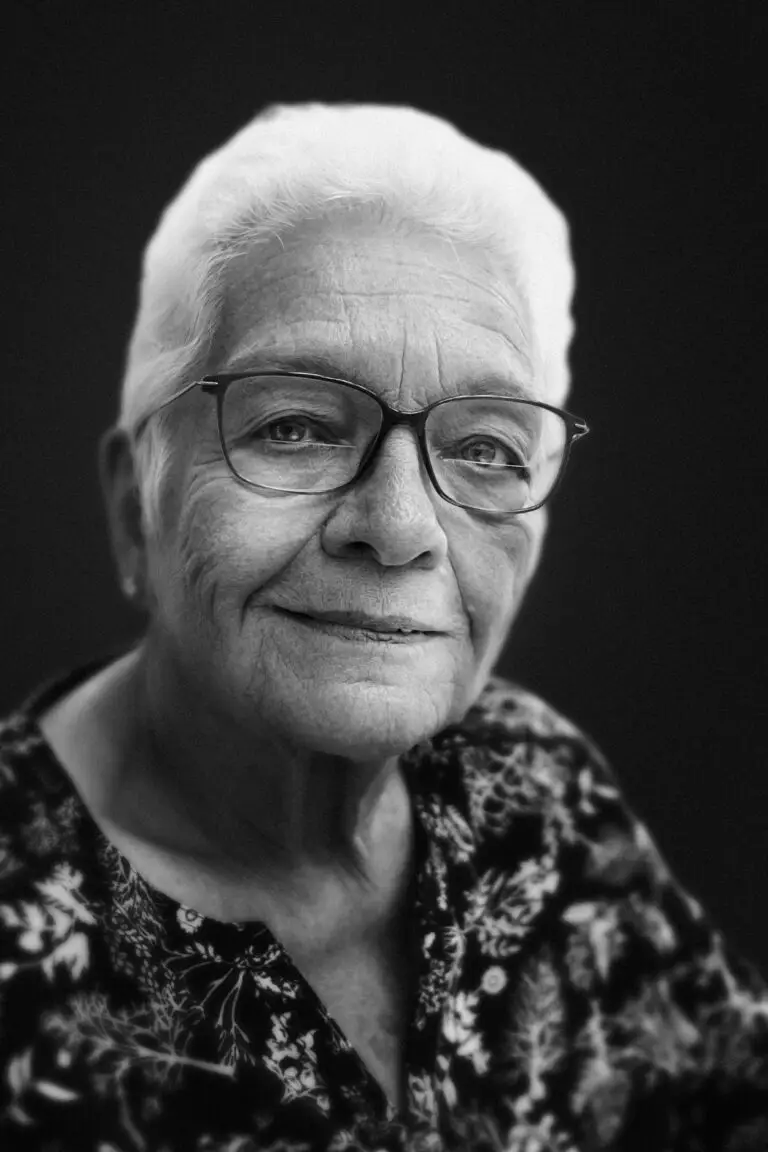 A black and white portrait of an older woman with short white hair, wearing glasses and a patterned blouse, looking calmly into the camera with a soft, knowing expression.