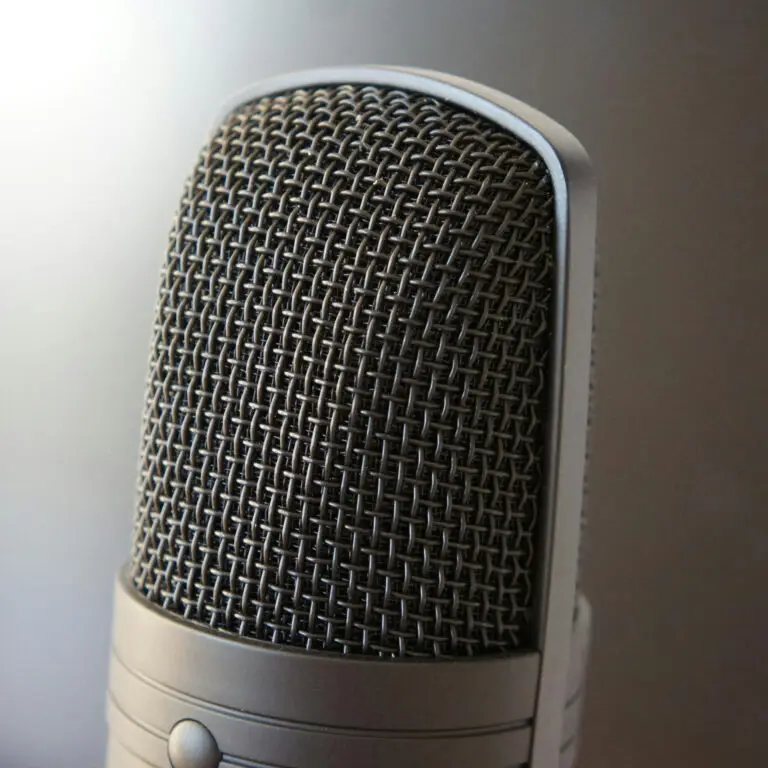 Close-up of a studio microphone with a metallic mesh grille and smooth metal body, set against a neutral background.