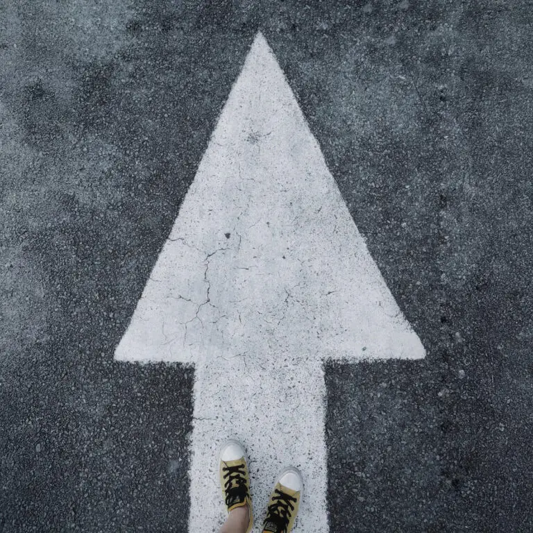 A large white arrow painted on asphalt, pointing forward, with a person wearing yellow sneakers standing at its base.