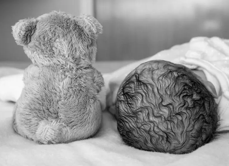 Black and white photo of a newborn baby lying next to a teddy bear, both seen from behind