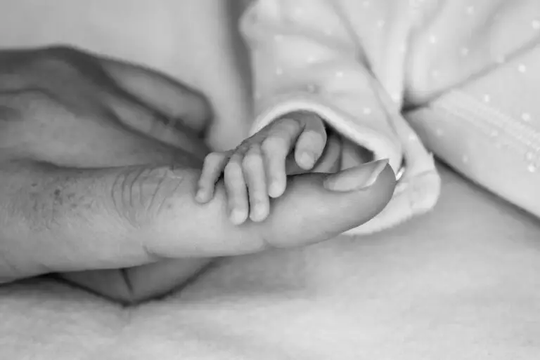 Black and white close-up photo of a newborn baby gently gripping an adult’s thumb with tiny fingers