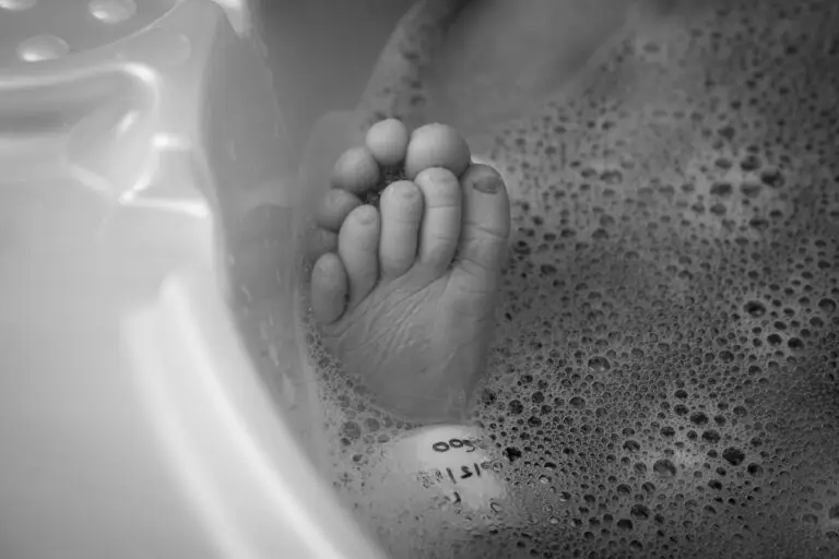 Black and white photograph of a newborn’s foot in a shallow bath, with hospital ID tag visible beneath soapy bubbles