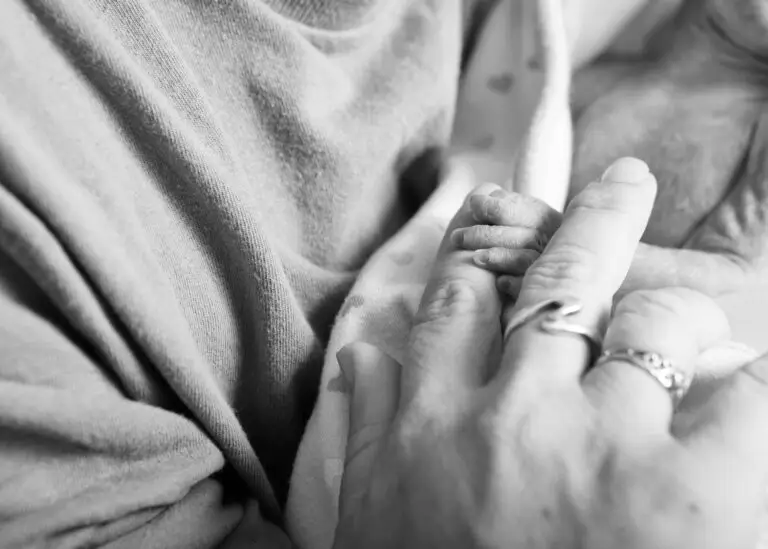 Black and white close-up of a newborn’s tiny hand gripping an adult’s finger, wrapped in a heart-patterned blanket
