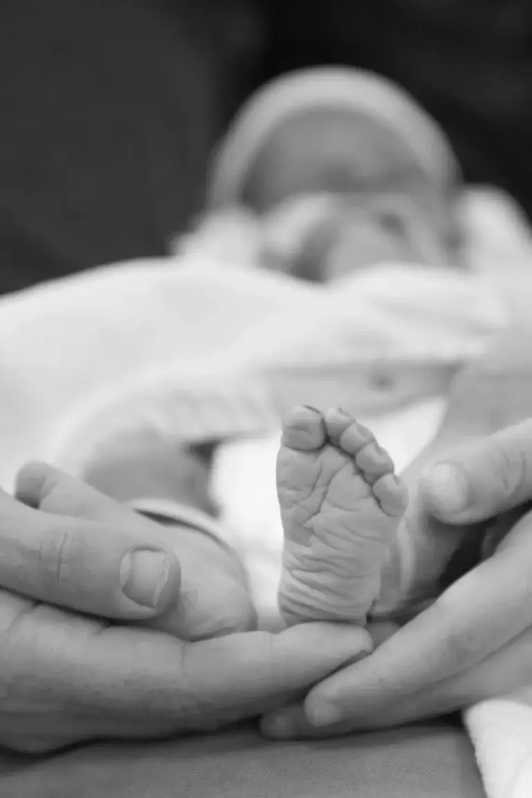 Black and white photo of a baby’s foot being gently supported by adult hands, with the rest of the baby softly blurred in the background