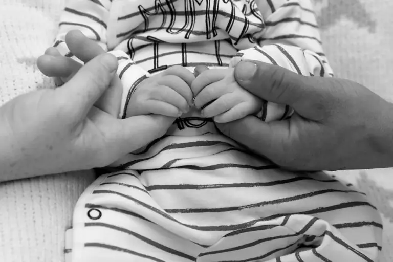Black and white photograph of a baby’s hands being gently held by two adult hands on either side