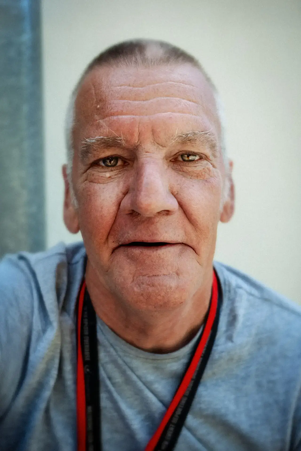 Colour close-up portrait of an older man in a grey T-shirt with a red-and-black lanyard, looking directly at the camera with a relaxed expression.